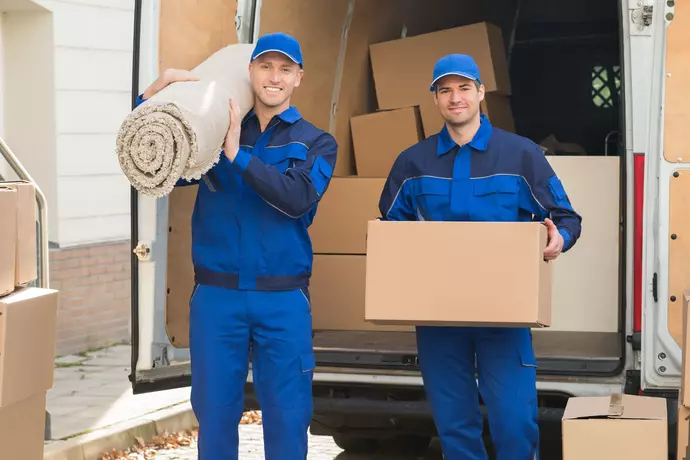 Zwei fröhliche Umzugshelfer in blauer Uniform stehen vor dem Lastwagen. Einer von ihnen hält einen aufgerollten Teppich, der andere einen Pappkarton in der Hand.